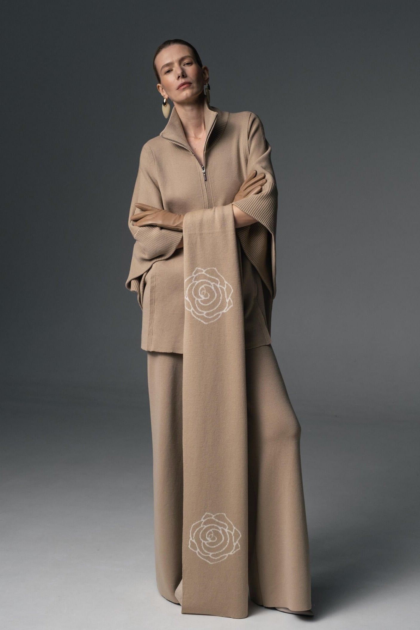 A female model wearing a beige three-piece wool outfit with a double-breasted coat, wide-leg trousers, and a scarf with rose embroidery.
