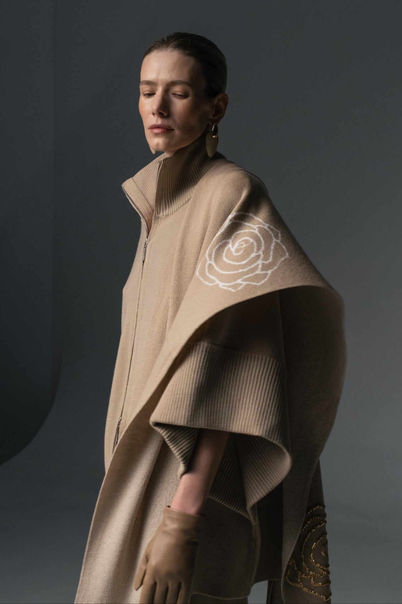 A person wearing a beige-colored merino wool cape with wide-leg trousers, both adorned with golden-thread rose embroidery.