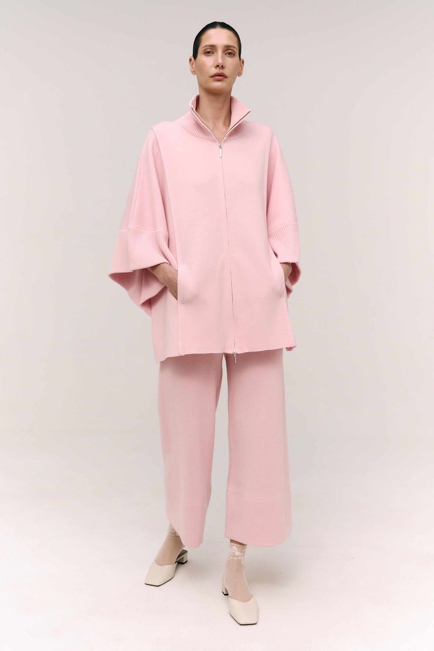 A model wearing a beige double-zip coat with a structured high collar and flowing sleeves, paired with matching cropped culottes. Footwear not shown.