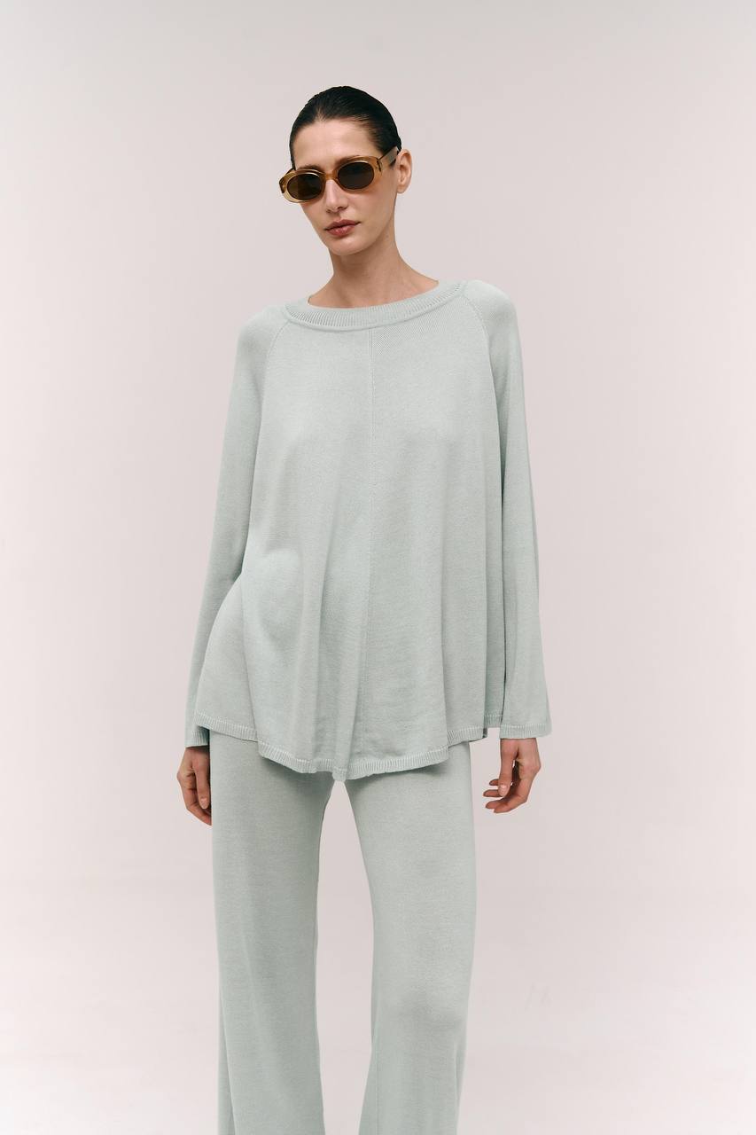 A model wearing a sage green cape-style top with raglan long sleeves and a wide crew neckline, paired with matching wide-leg trousers.
