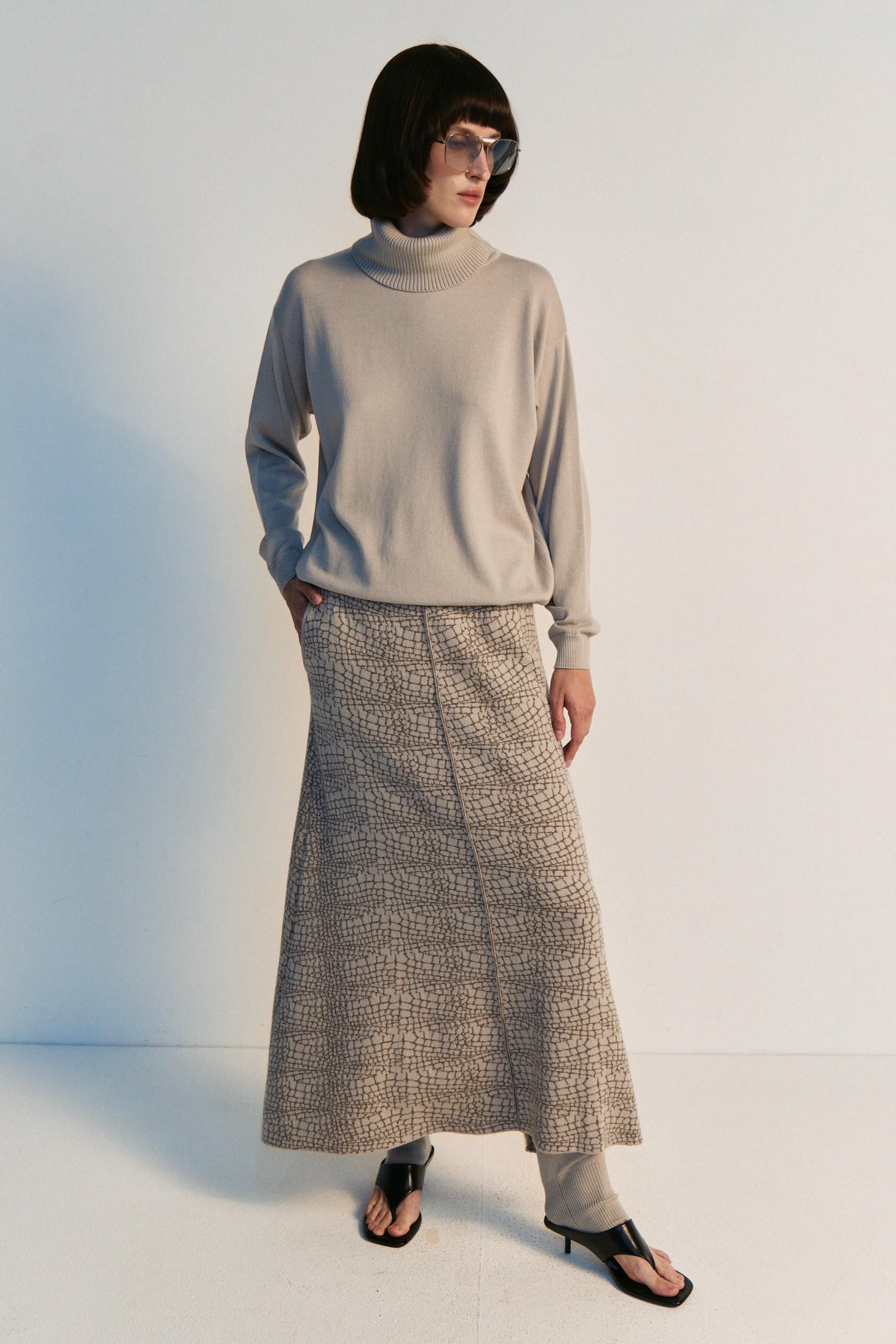 Person wearing a light gray turtleneck sweater and patterned skirt on a white background