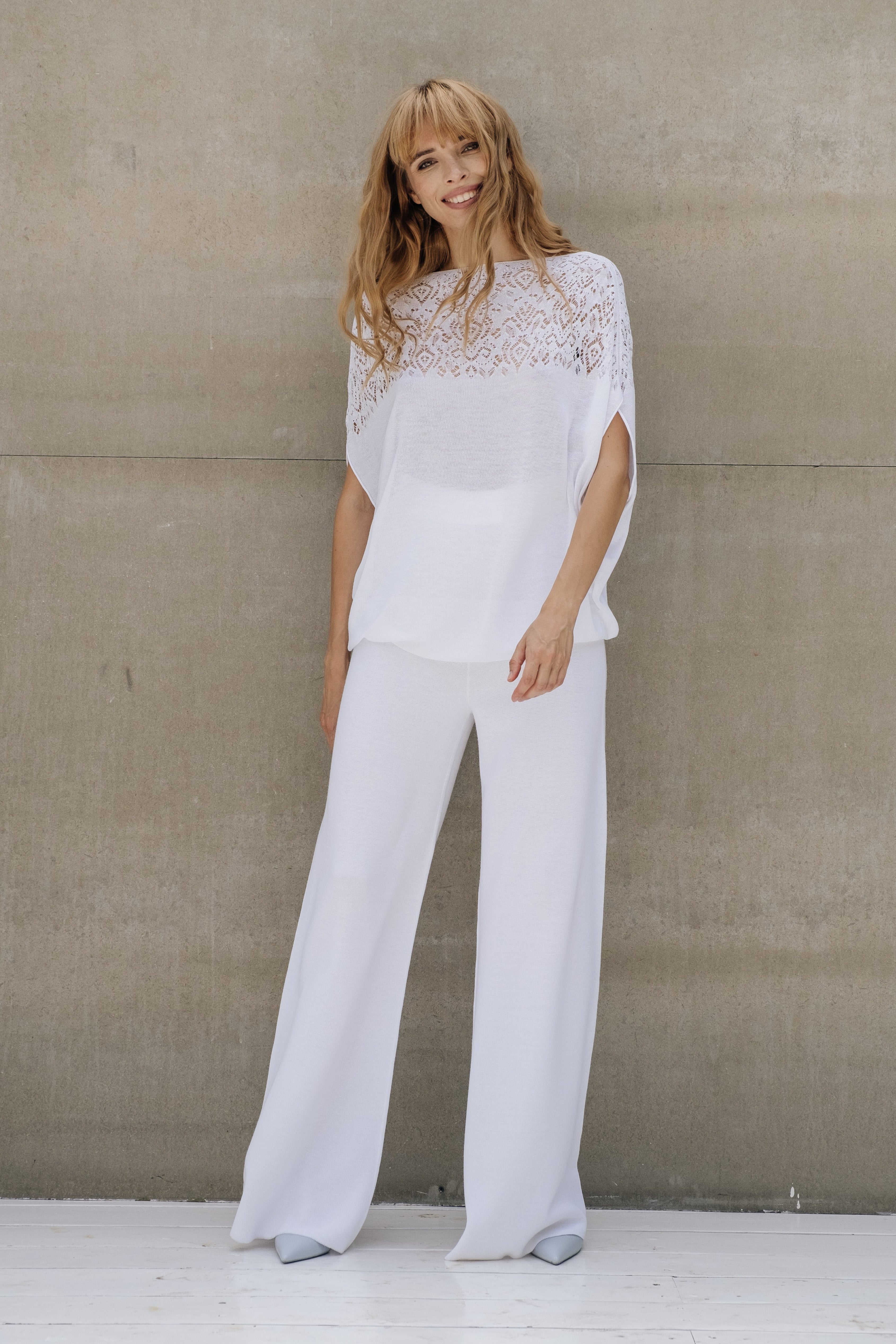 A woman is posing wearing a white crocheted top with a wide neckline and a pair of white trousers. She is also wearing sunglasses and open-toe shoes.