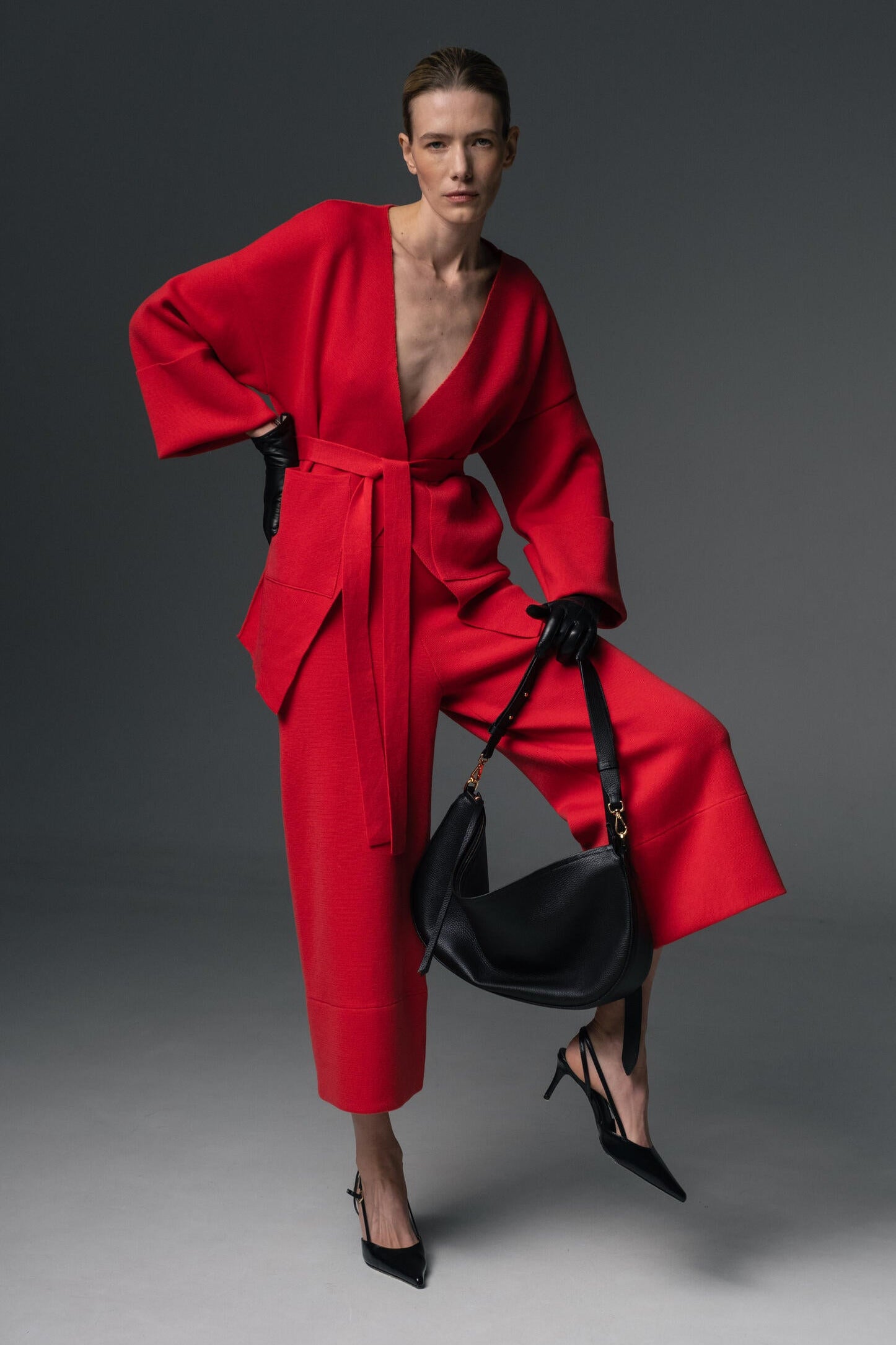 Look 49 – Second photo of a Model in red merino wool kimono jacket with belt and matching culottes, styled with black gloves, heels, and clutch against a grey studio backdrop.