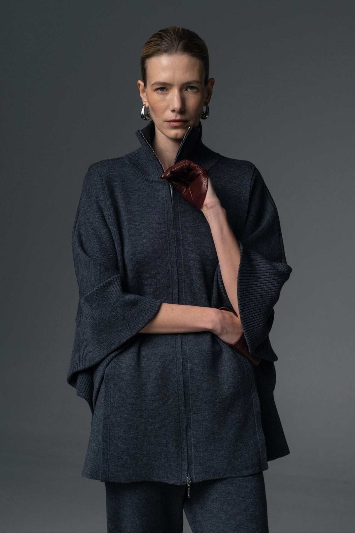 A model wearing a double zip-up coat with a high collar and tapered knit trousers, holding a red accessory.