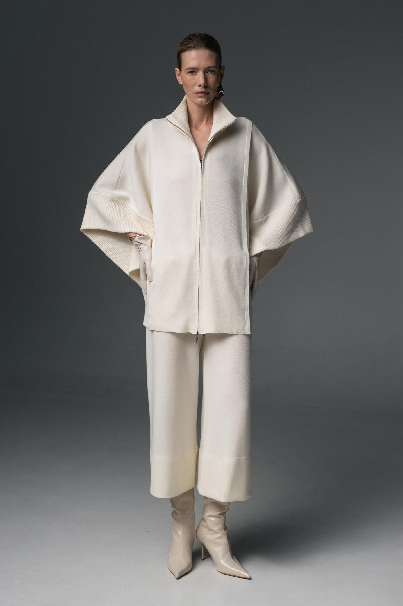 A model wearing a beige double-zip coat with a structured high collar and flowing sleeves, paired with matching cropped culottes. Footwear not shown.