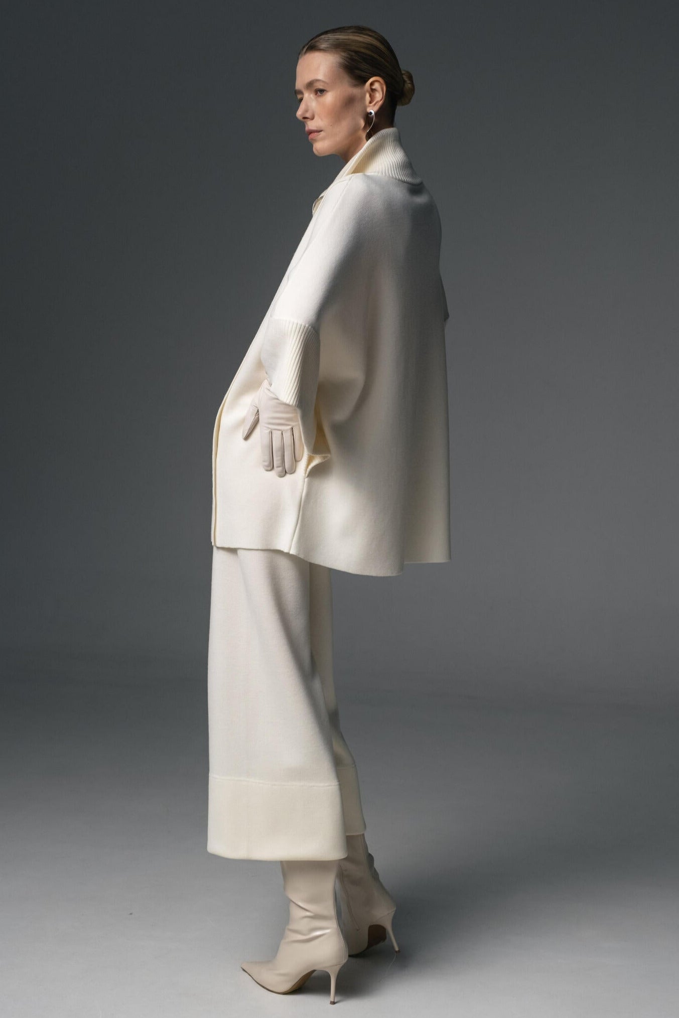 A model wearing a beige double-zip coat with a structured high collar and flowing sleeves, paired with matching cropped culottes, and high-heeled boots.