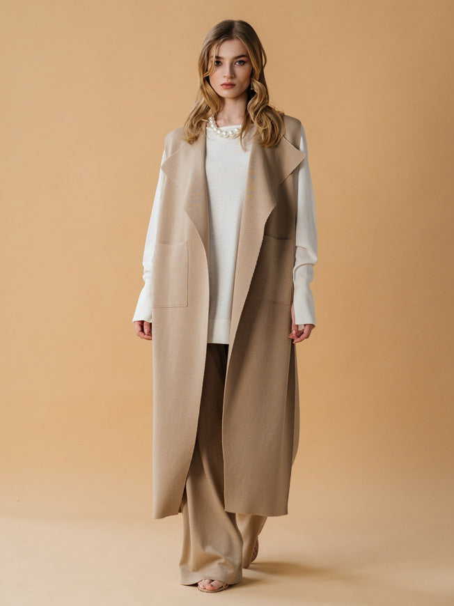 A female model wearing a beige sleeveless merino wool vest with sculptural lapels and a self-tie belt, paired with a white top and dark pants.