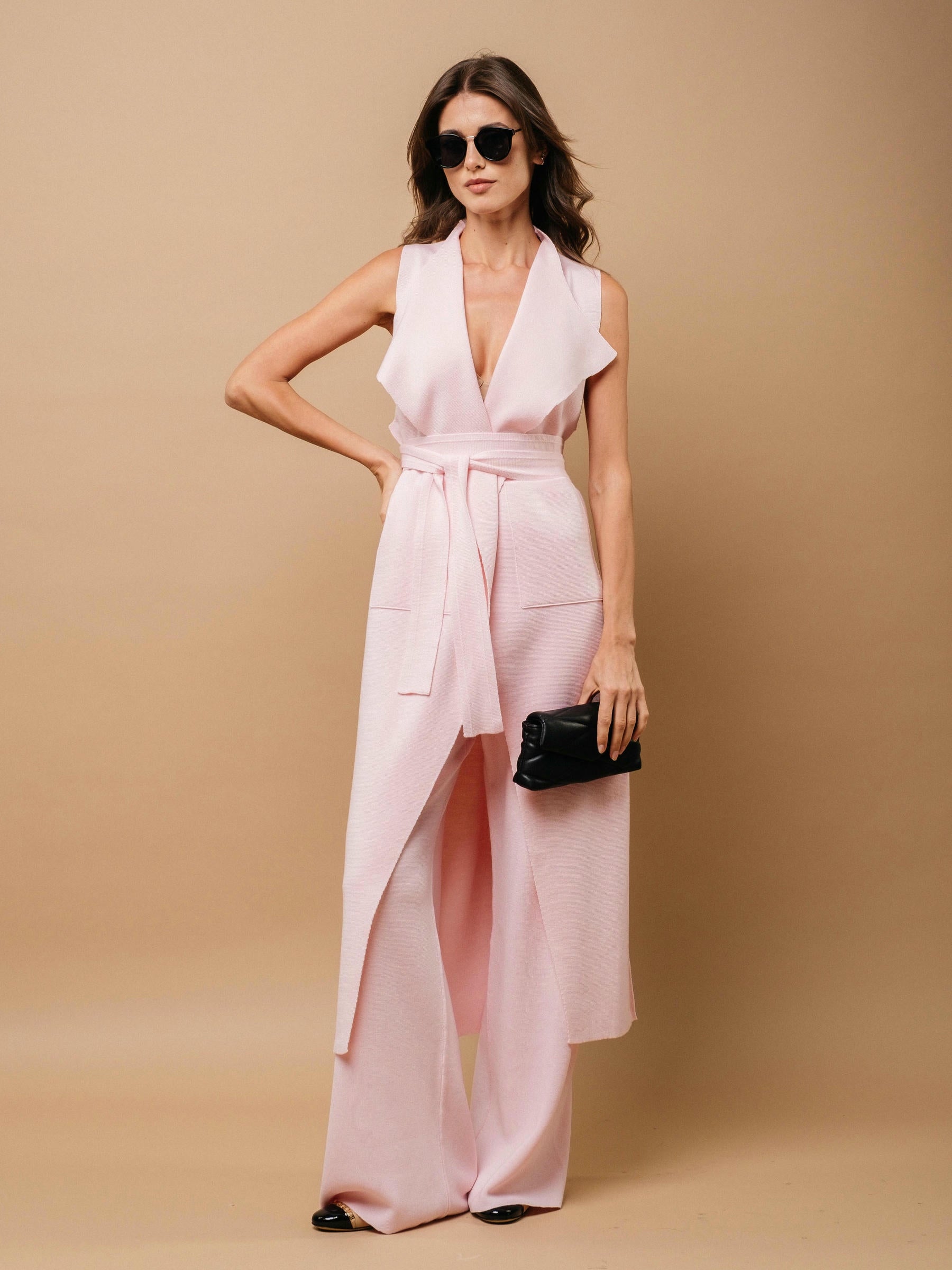 Woman in a light pink vest and pants with a black clutch against a beige background.