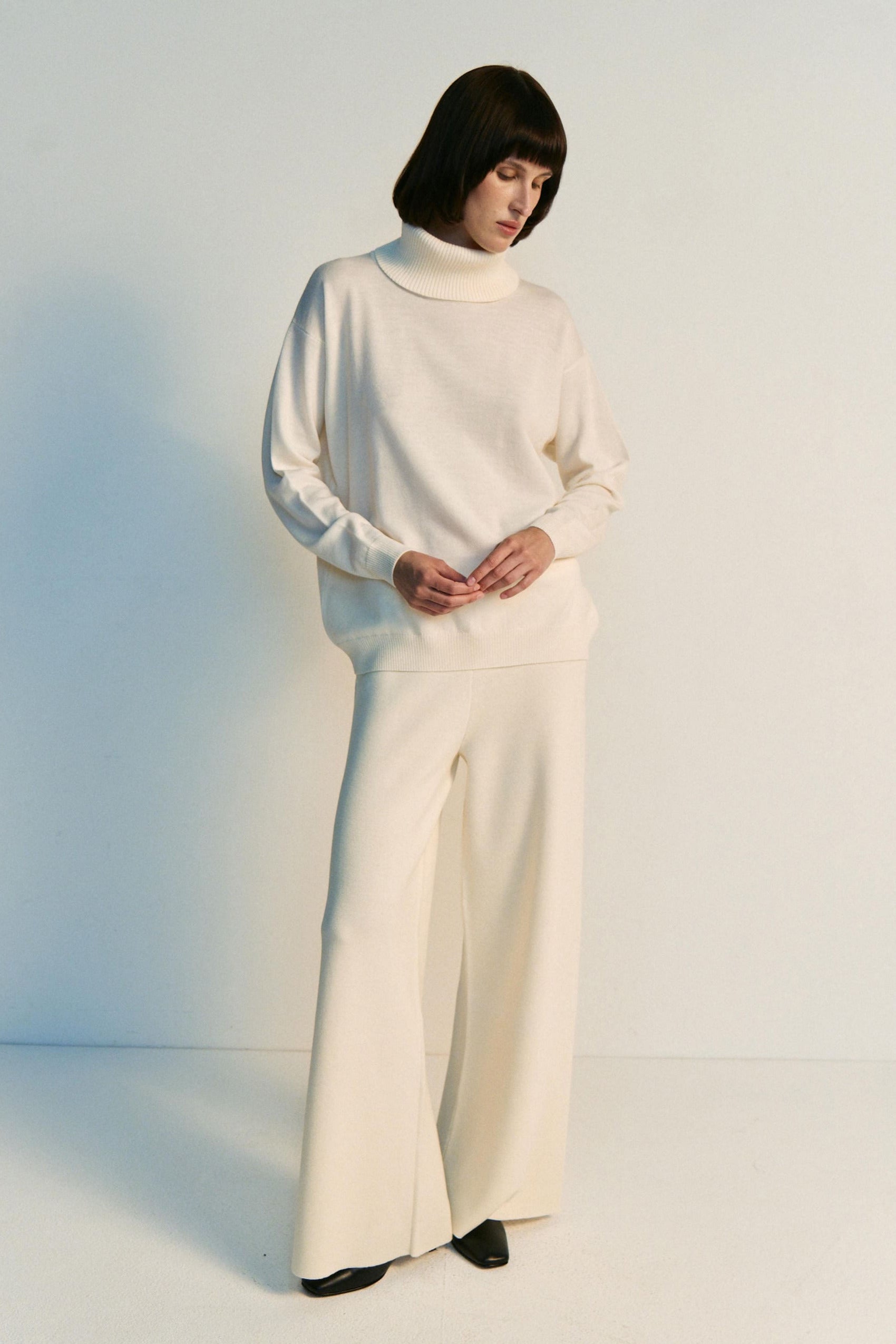 Person wearing a white turtleneck and wide-leg pants against a plain background