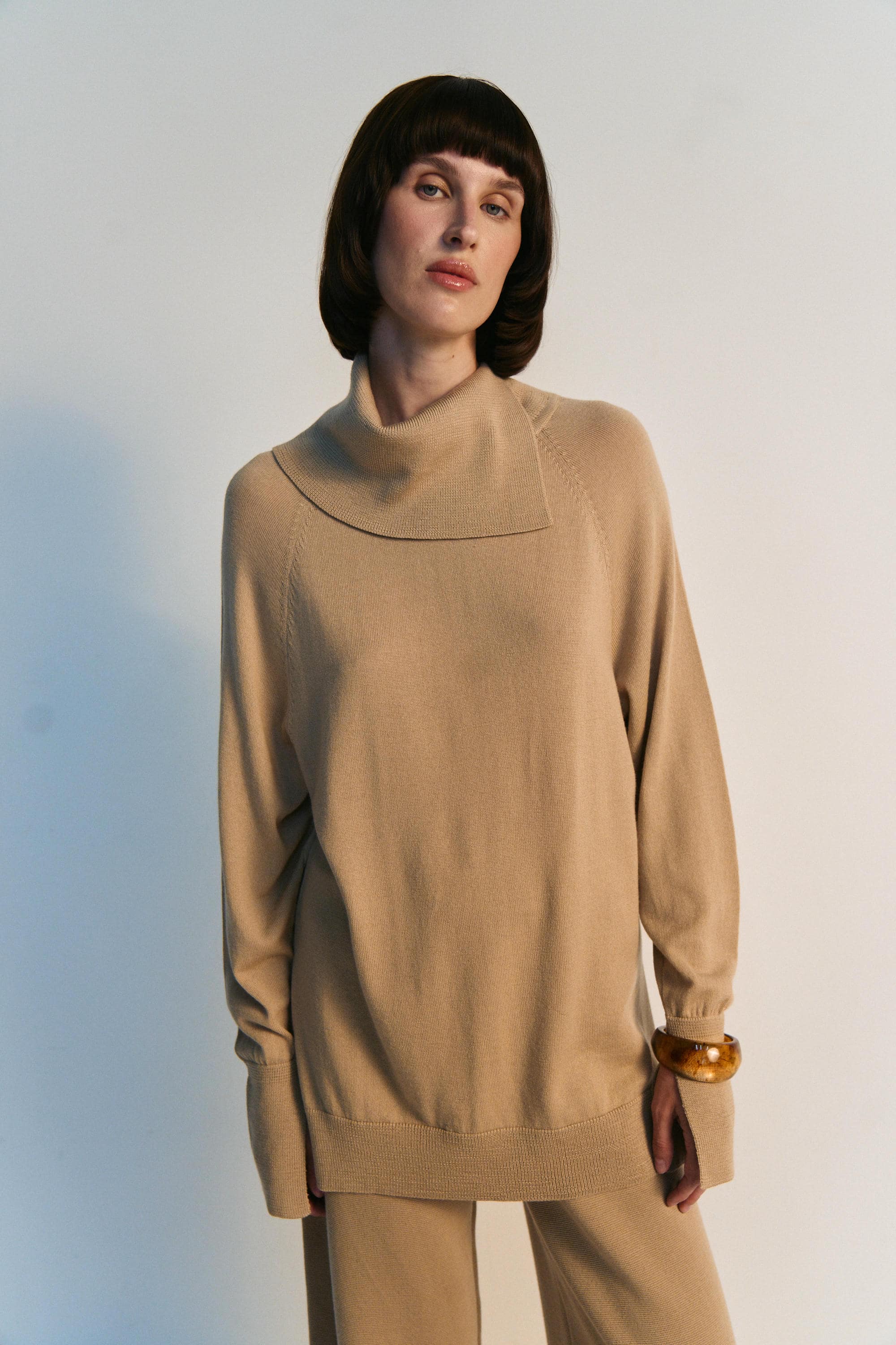 Woman wearing a beige sweater and pants against a light background