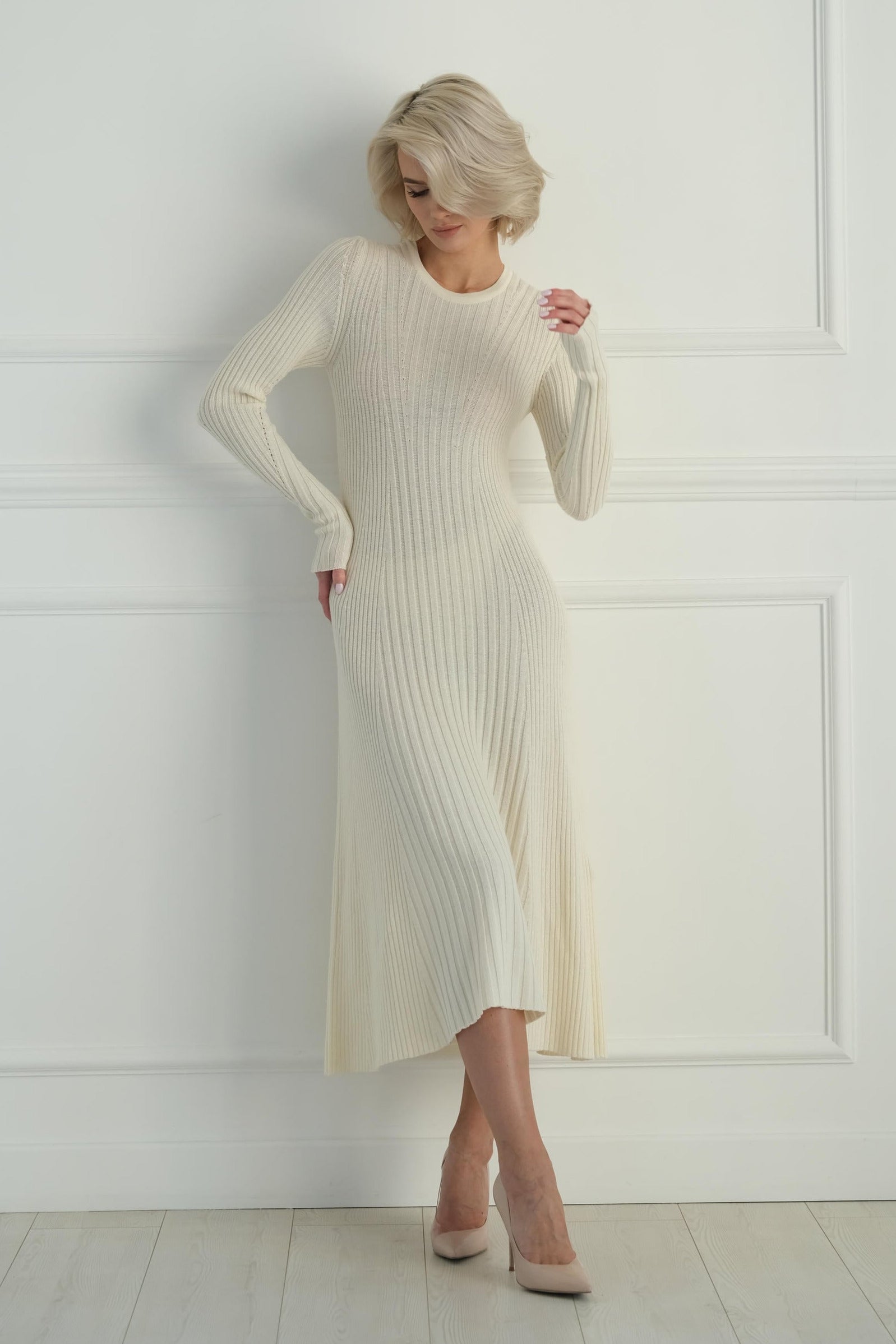 Woman wearing a long white ribbed dress against a white wall.