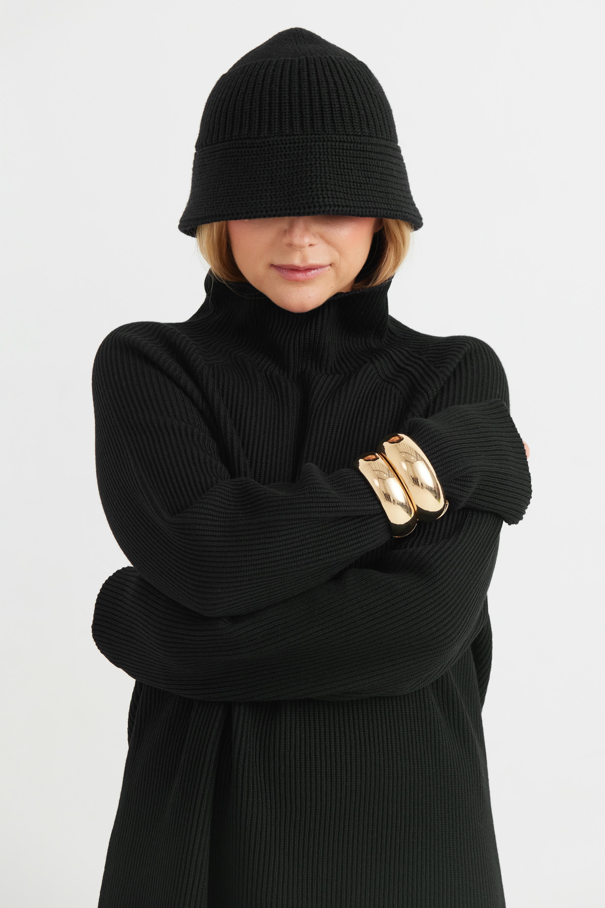 Person wearing a black knit hat and black sweater with gold arm bands on a white background