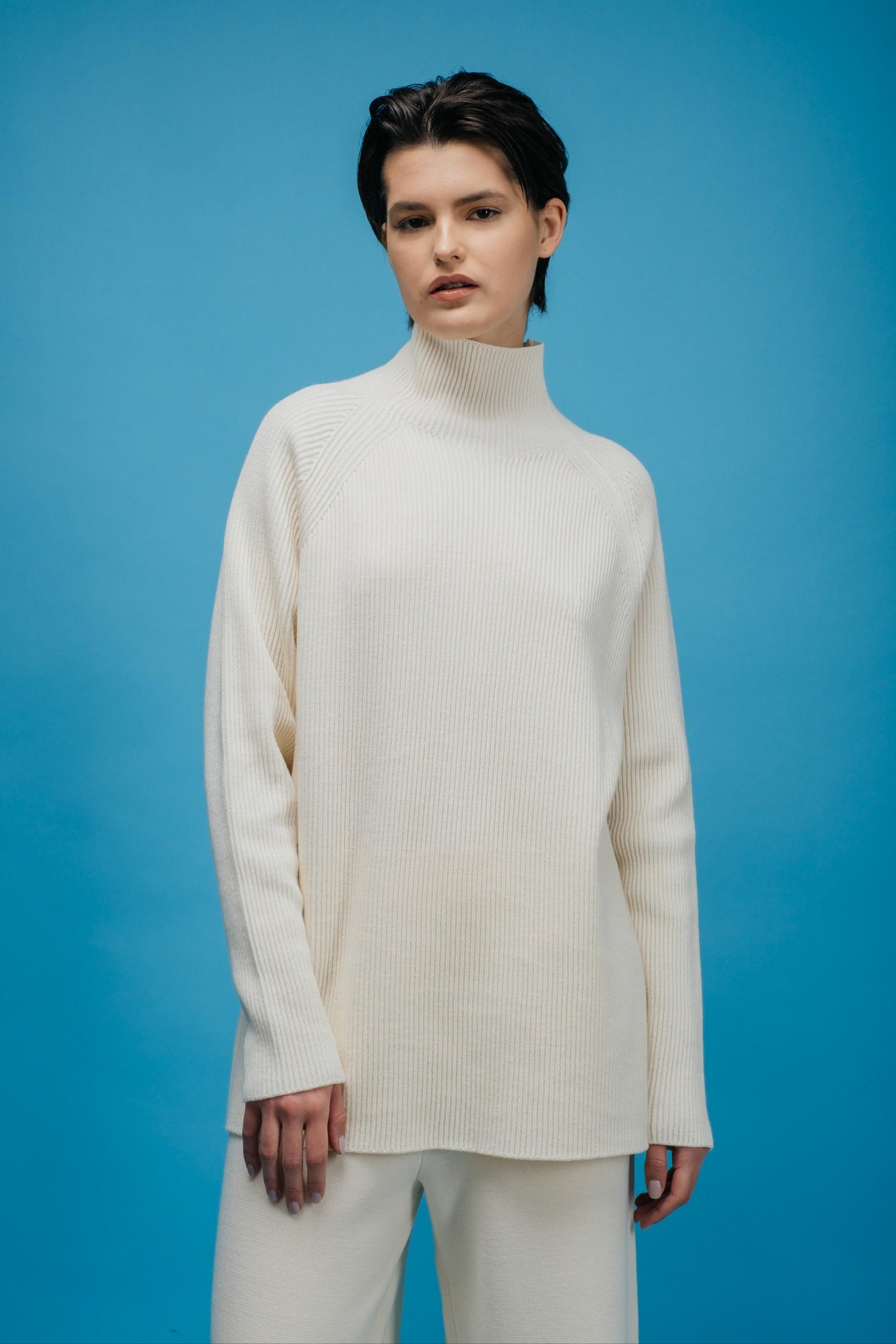 Person wearing a white turtleneck sweater and pants against a blue background