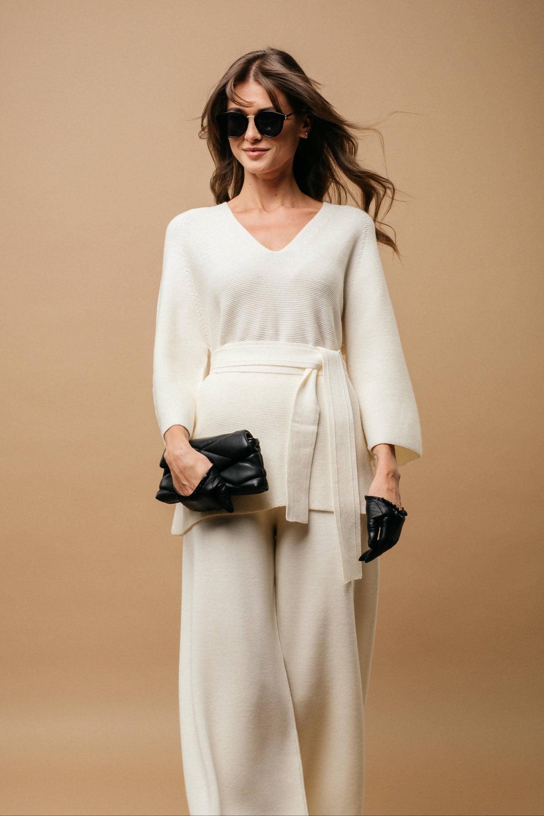 Woman in a white outfit holding black handbags against a beige background