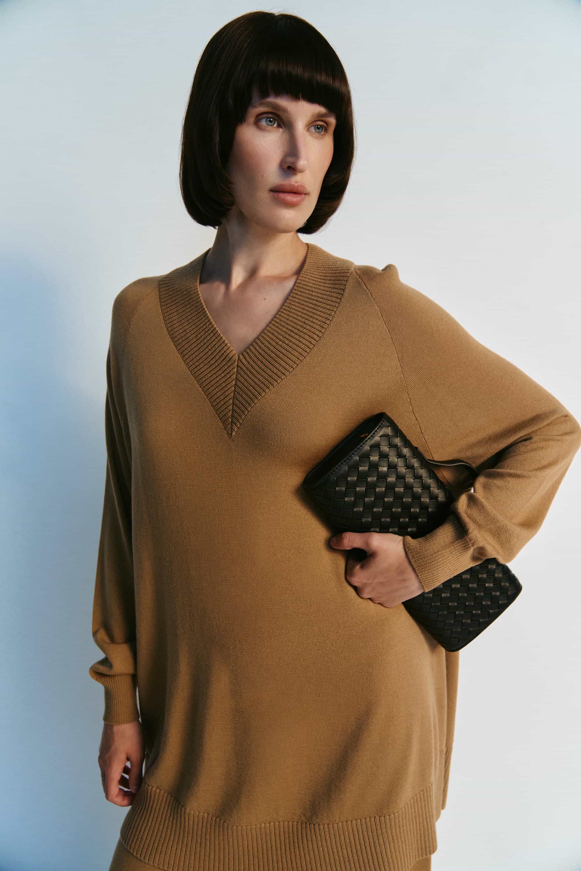 Woman wearing a brown sweater dress holding a black clutch against a light blue background