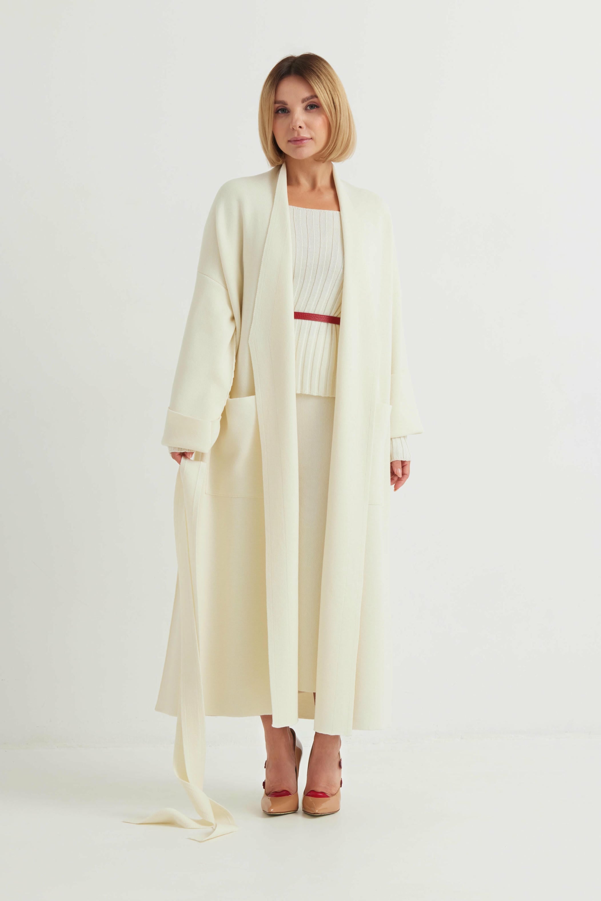 Woman wearing a long cream coat over a white outfit with a red belt on a white background