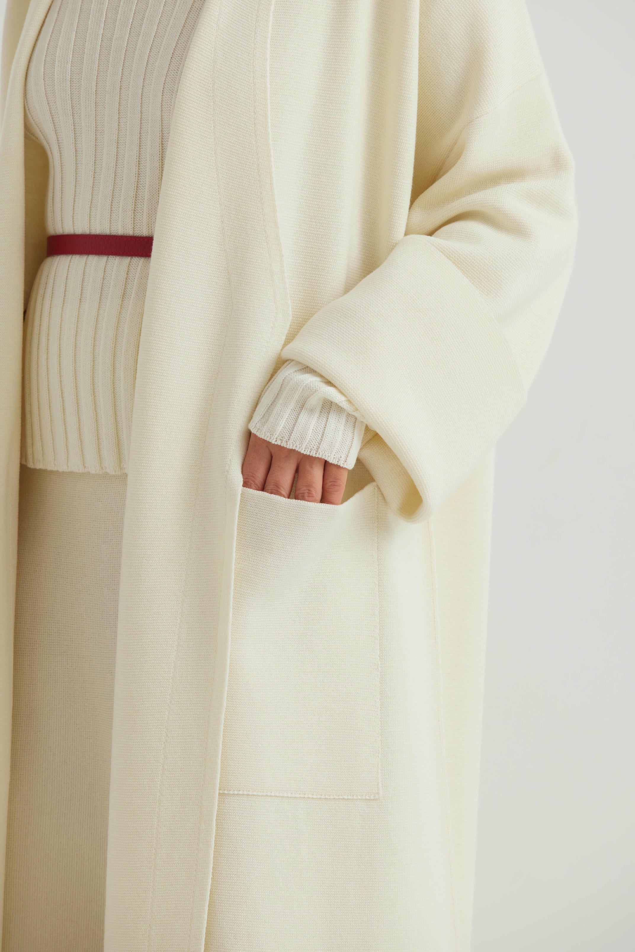 Close-up of a person wearing a cream coat with a white ribbed sweater underneath on a light gray background.