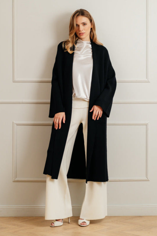 Woman wearing a black coat over a white outfit against a light-colored wall.