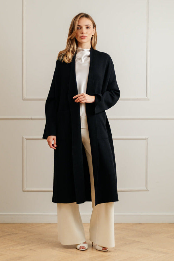 Woman wearing a black coat over a white outfit against a light-colored wall.