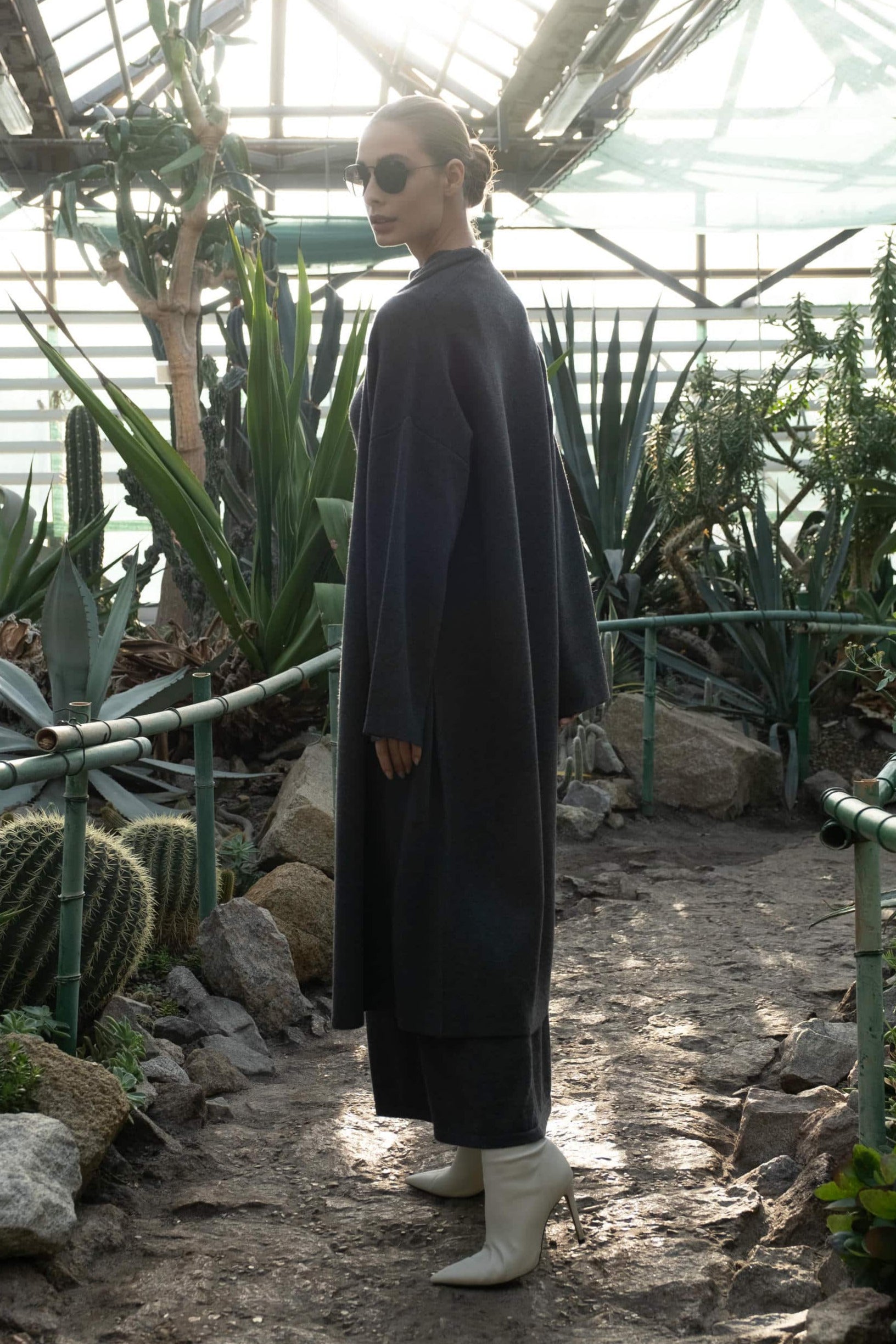 Person in a long coat standing in a greenhouse with plants and rocks.