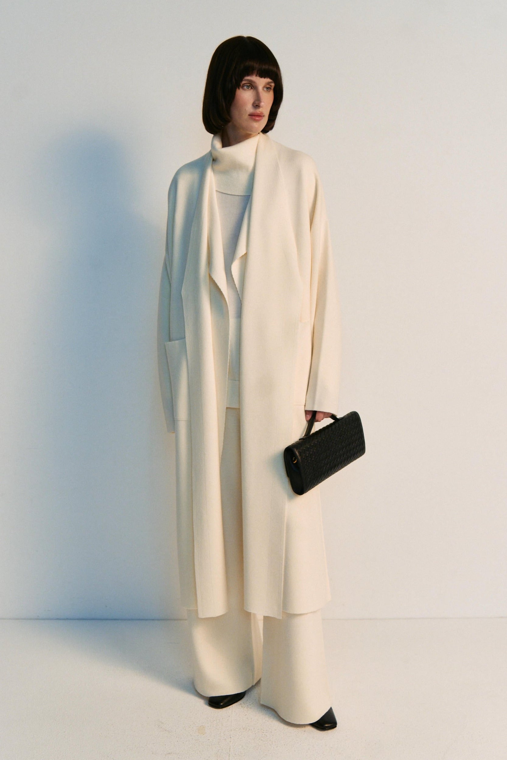 Person wearing a long cream coat and holding a black clutch against a white background