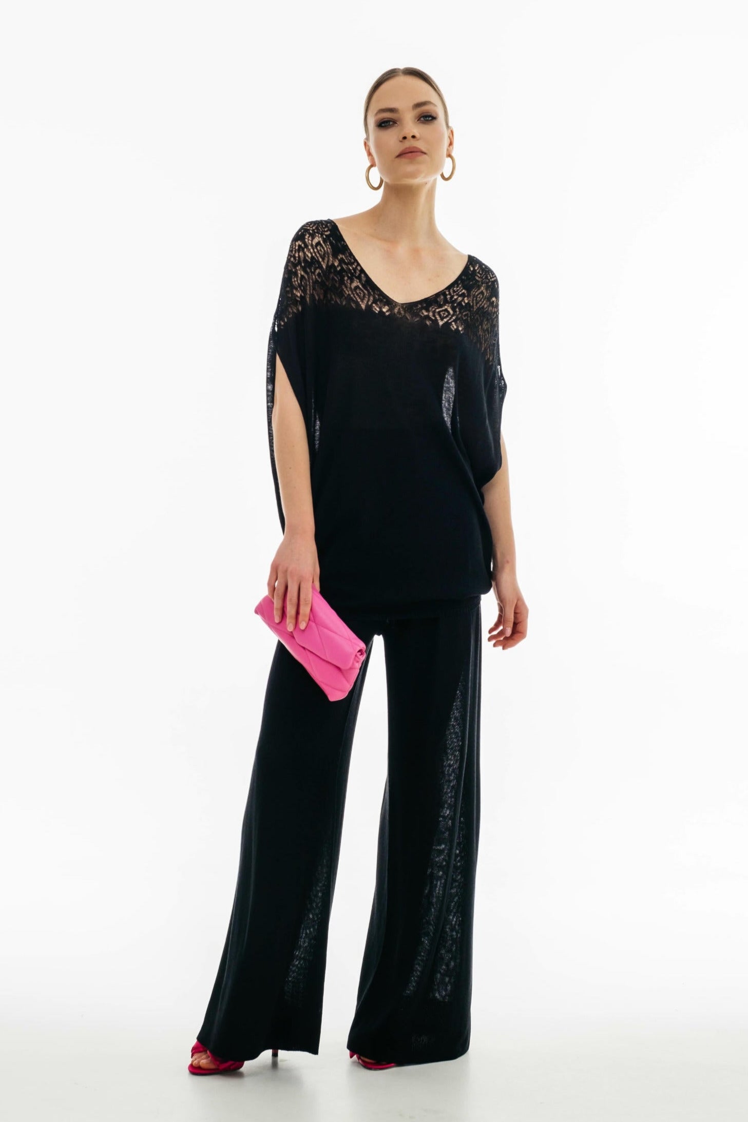 Woman wearing a black lace top and wide-leg pants holding a pink clutch on a white background