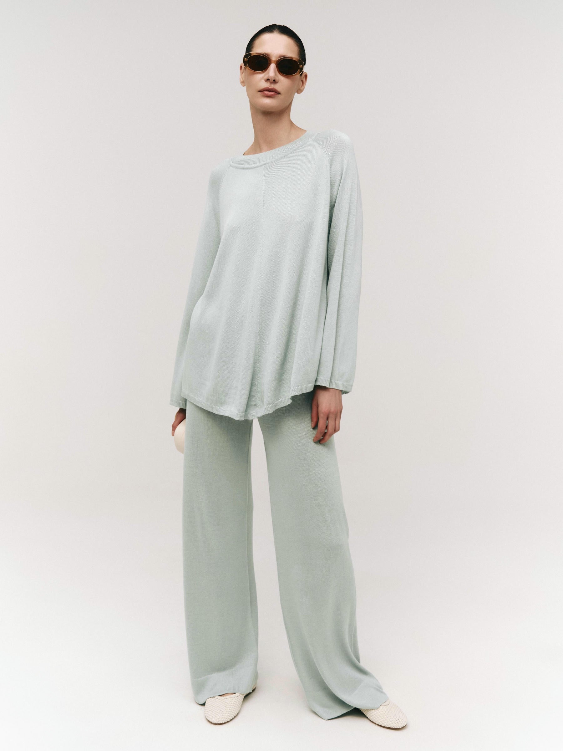 A model wearing a sage green cape-style top with raglan long sleeves and a wide crew neckline, paired with matching wide-leg trousers.