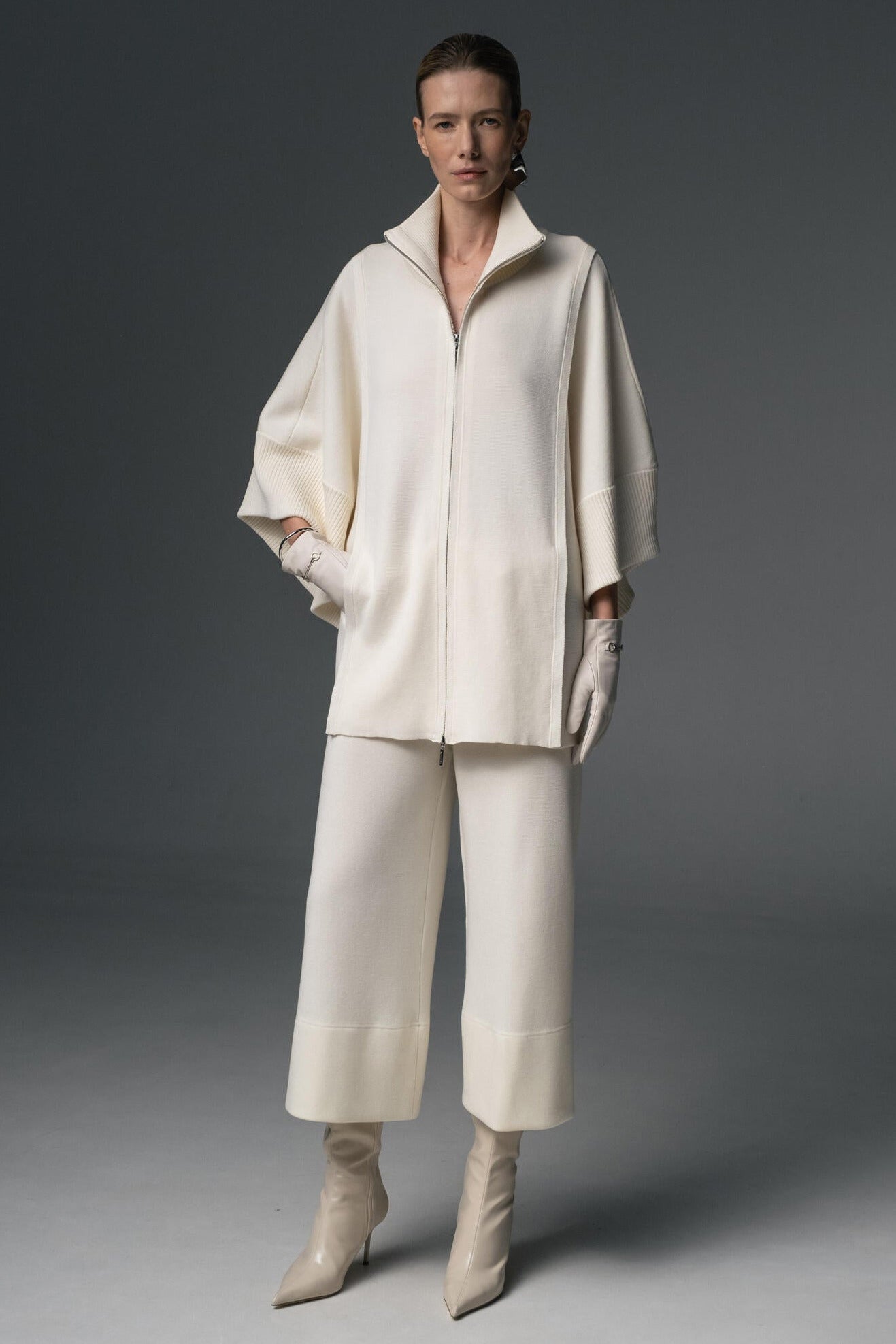 A model wearing a beige double-zip coat with a structured high collar and flowing sleeves, paired with matching cropped culottes. Footwear not shown.
