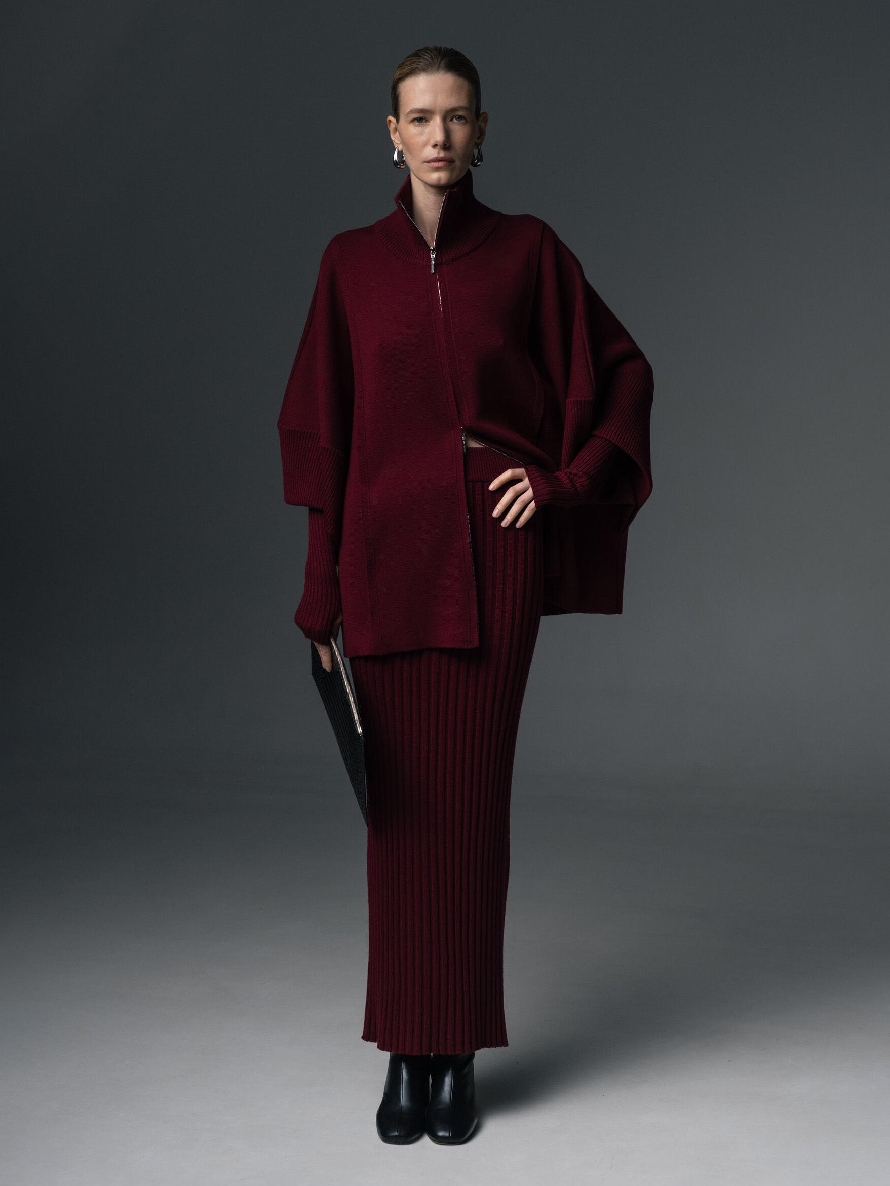 Model wearing a burgundy merino wool ribbed maxi skirt and oversized zip jacket, styled with black boots and silver earrings against a dark grey background.