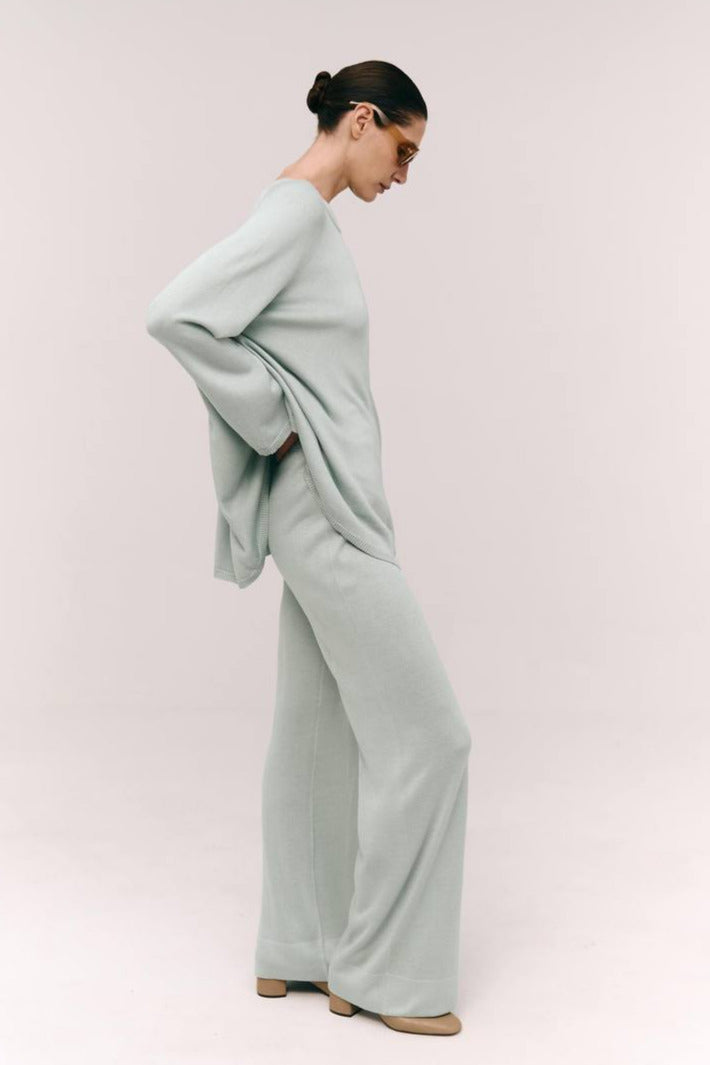 A model wearing a minimalist cape-style top in a cool sage color, with raglan long sleeves and a wide crew neckline.