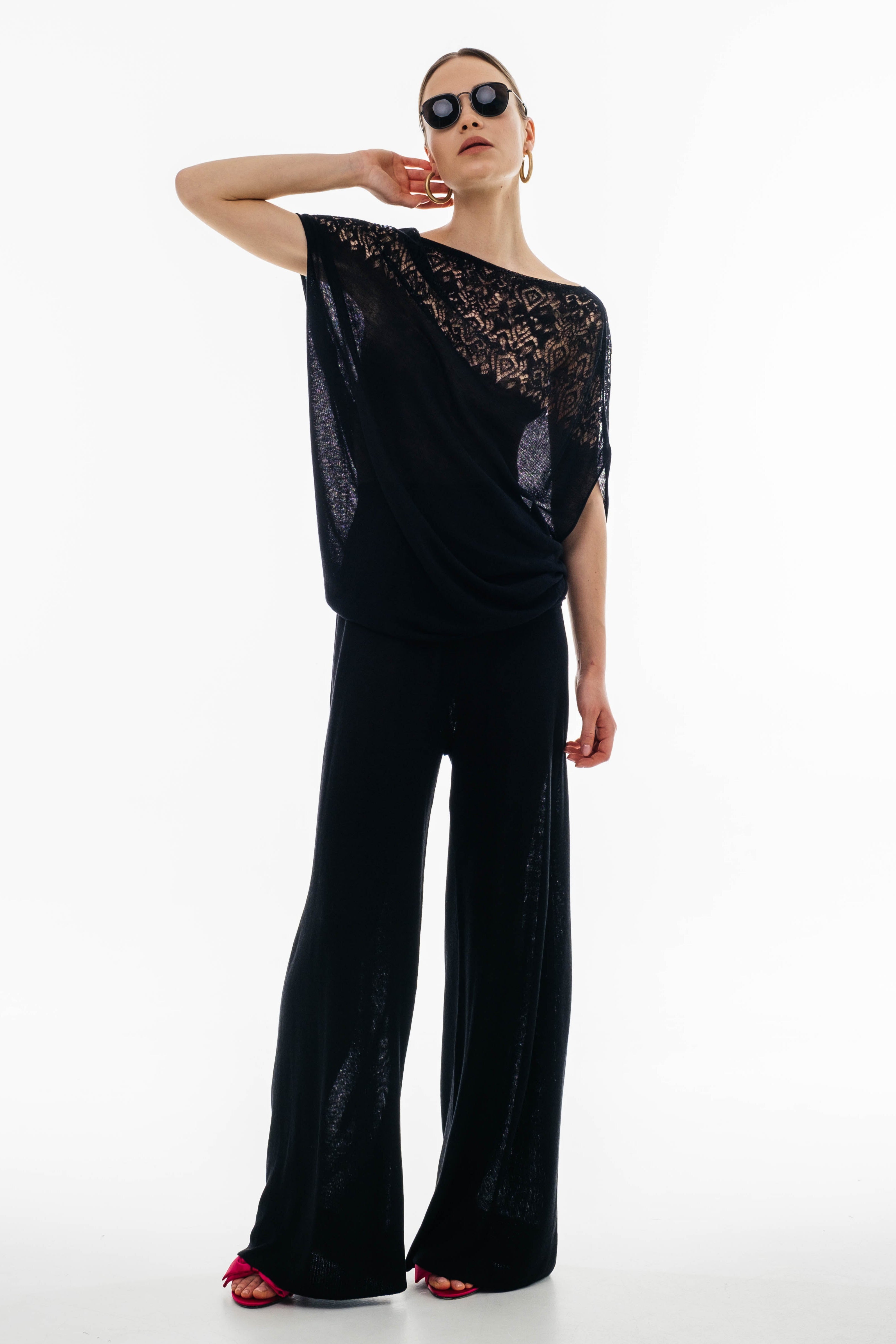 A woman is posing wearing a black crocheted top with a wide neckline and a pair of black trousers. She is also wearing sunglasses and open-toe shoes.