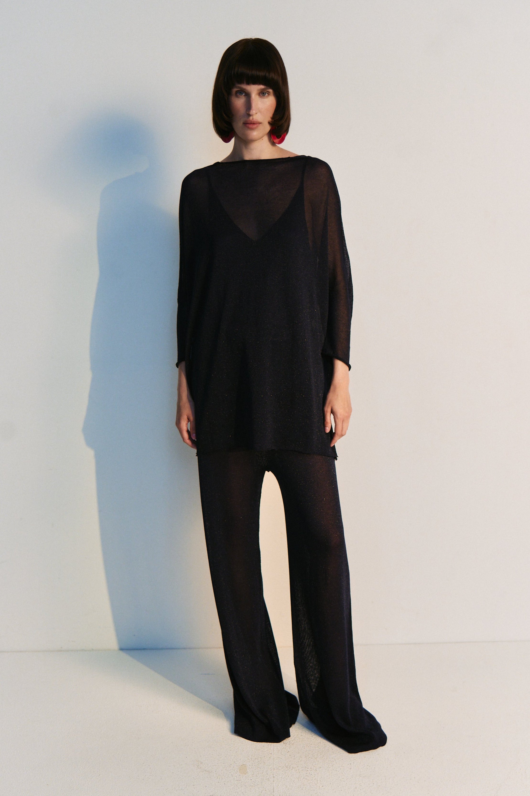 Person wearing a black outfit standing against a white wall with a blue shadow effect.