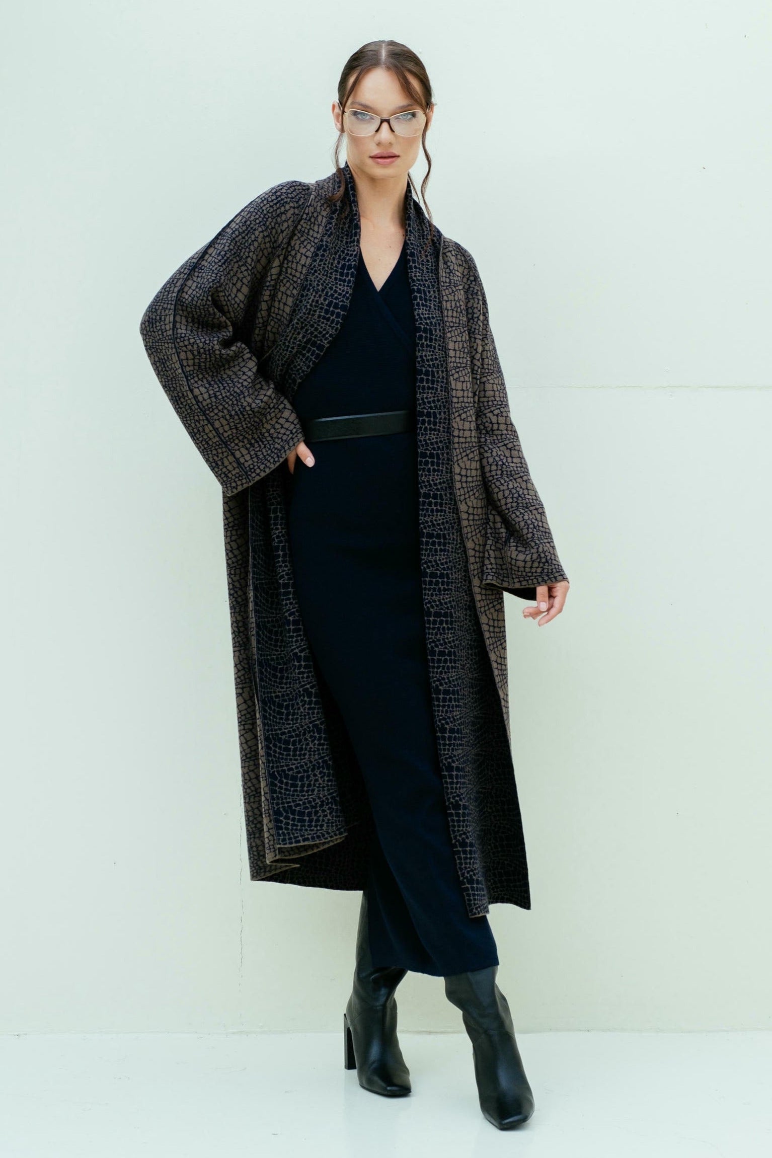 A female model wearing a long belted coat with a textured jacquard weave, in a patterned color with python-inspired print, paired with a black top and boots.