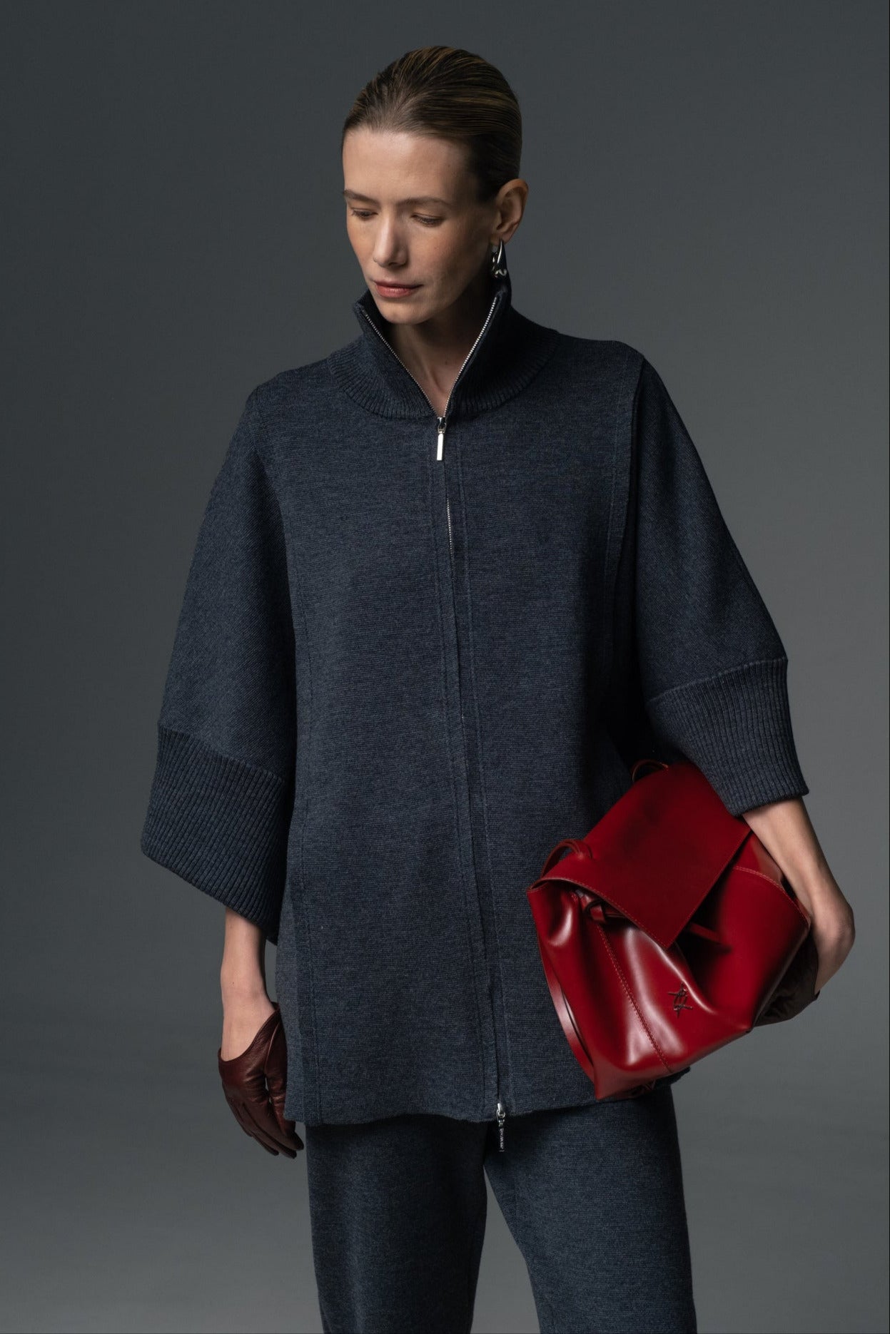 A person wearing a navy-colored double zip-up coat with a high collar and tapered knit trousers, holding a red bag.
