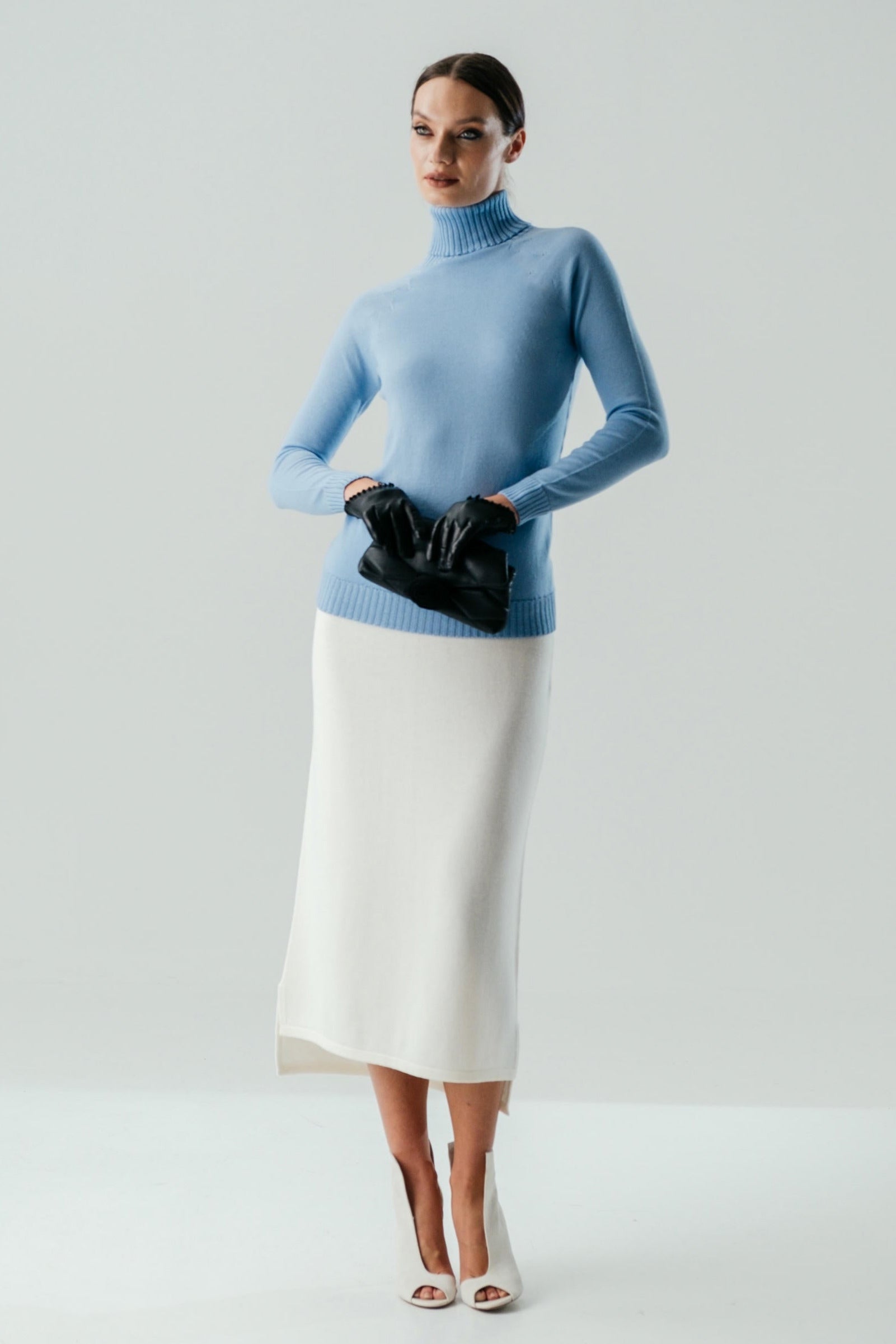 A woman wearing a blue turtleneck sweater and a white skirt. She is holding a black accessory, possibly a clutch.