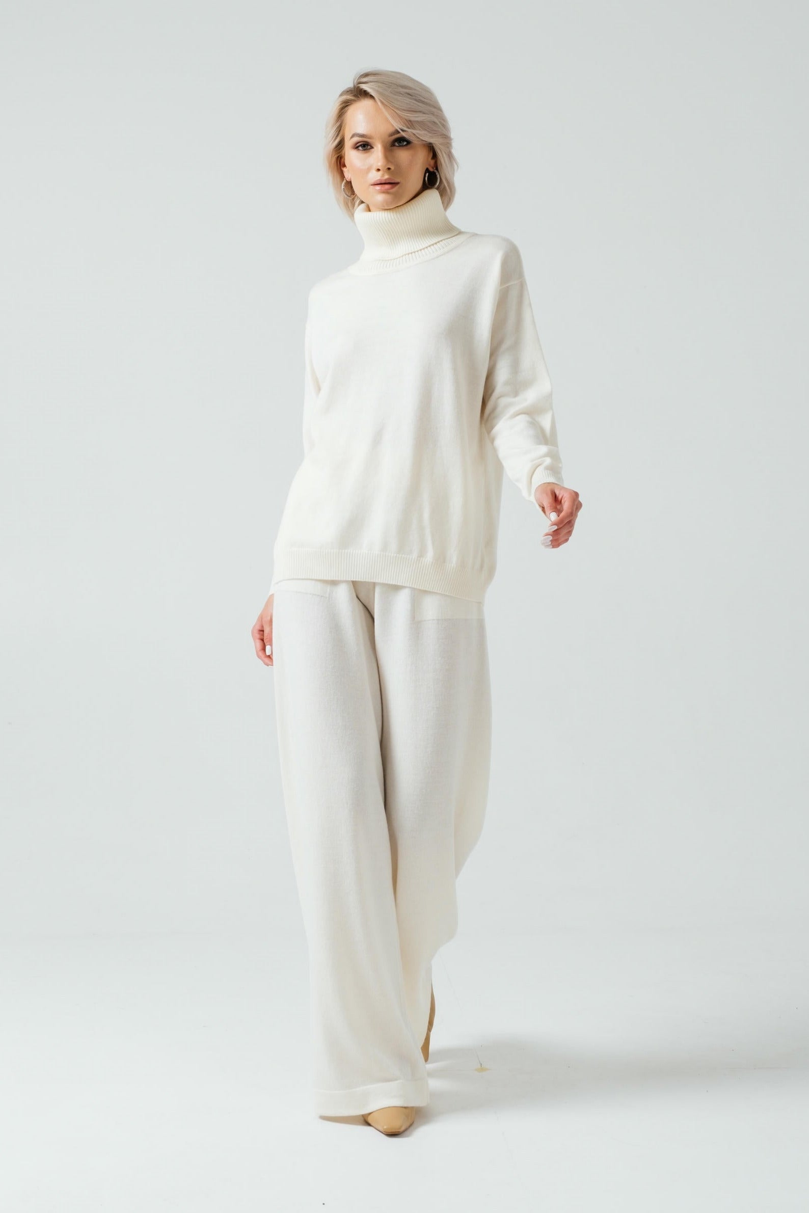Woman wearing a white turtleneck sweater and beige pants on a plain background