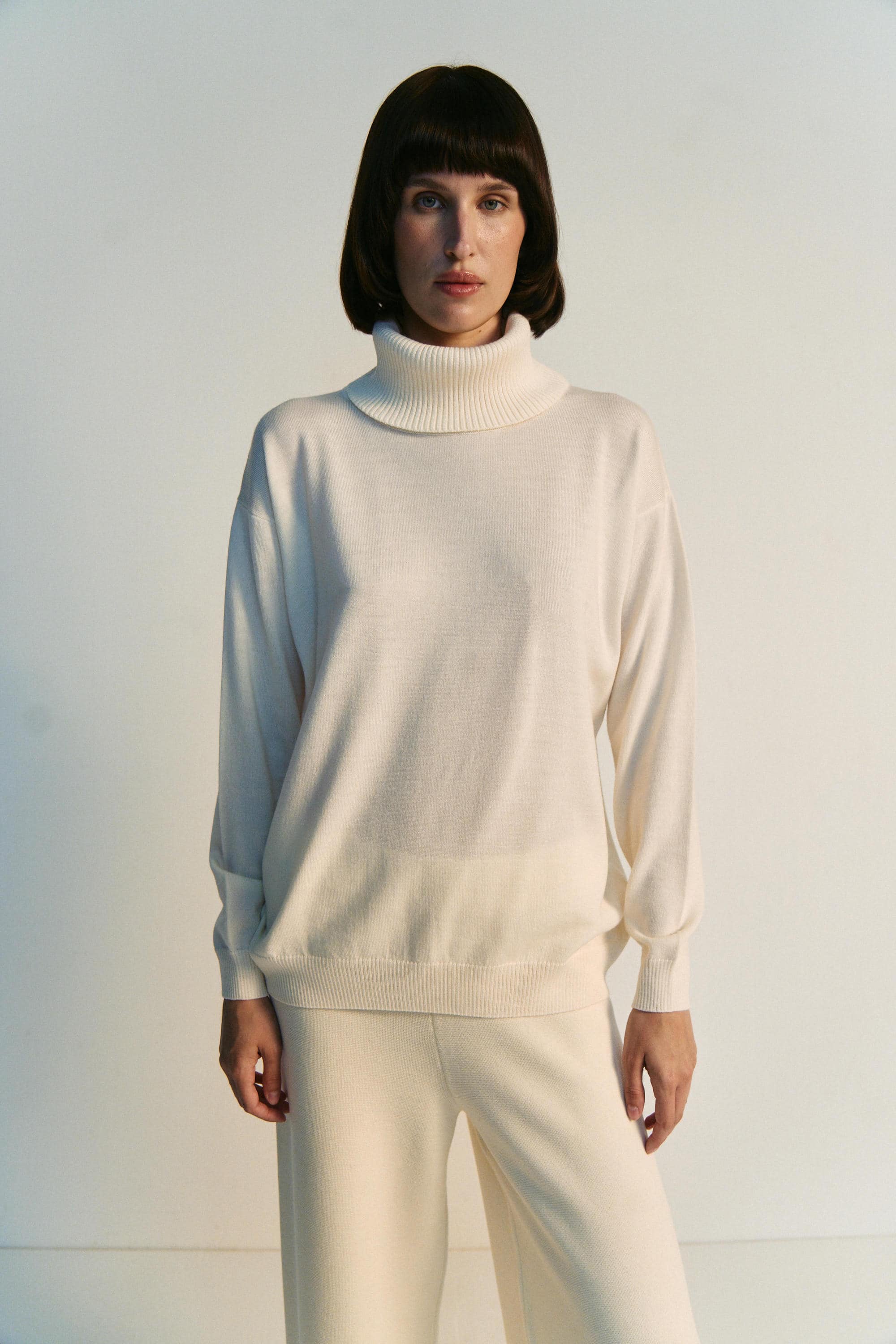 Woman wearing a white turtleneck sweater and pants against a plain background