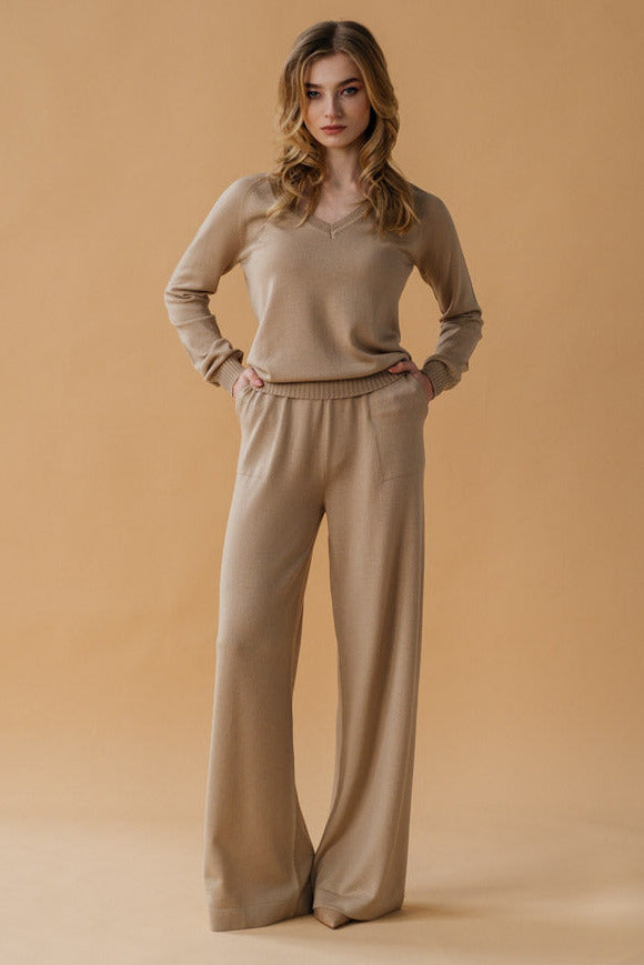 A female model standing in a greenhouse wearing wide-leg beige palazzo pants with a matching top, paired with a blue scarf.