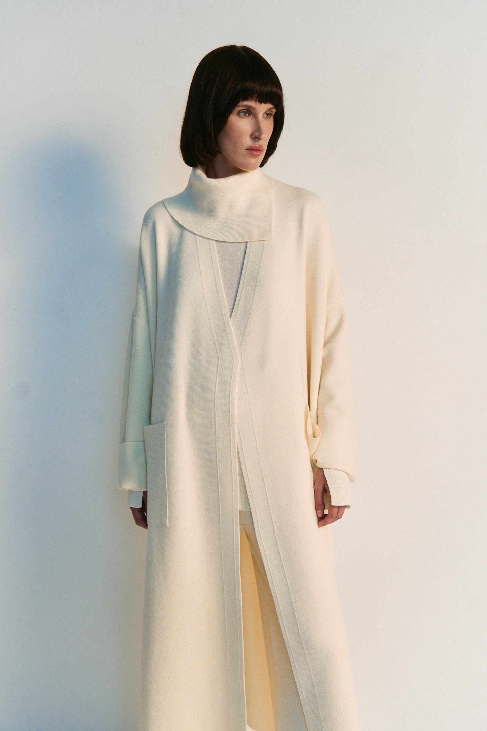 Person wearing a long, light-colored coat against a plain background