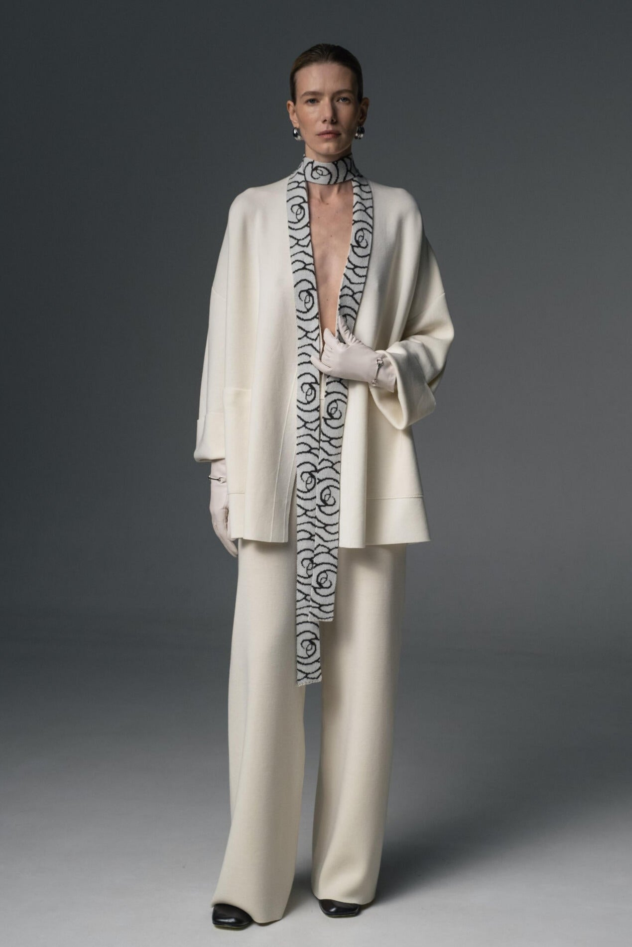 A model wearing a white coat with a geometric pattern on the collar, paired with matching trousers and a narrow scarf.