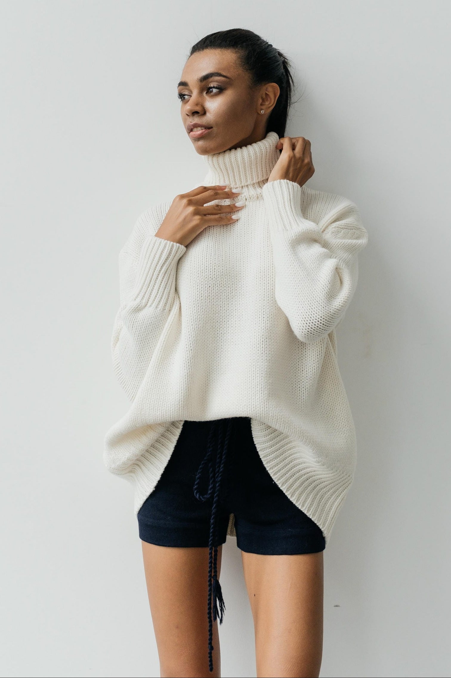 Woman wearing a white turtleneck sweater and black shorts against a plain background